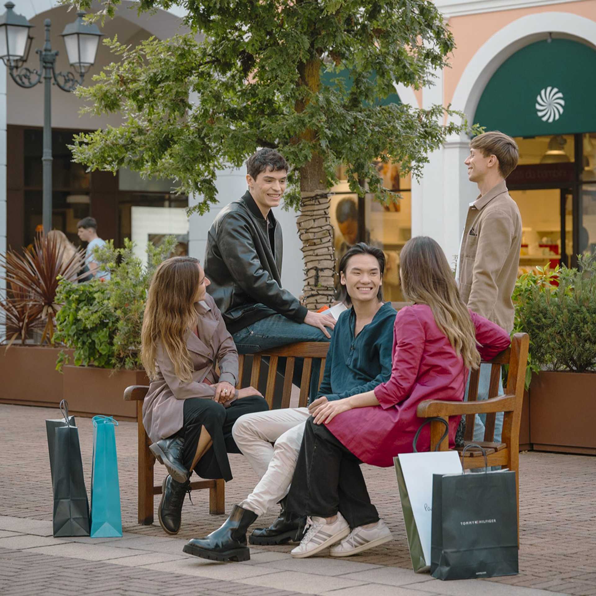 Group of friends talking on a bench at Noventa di Piave Designer Outlet with shopping bags.
