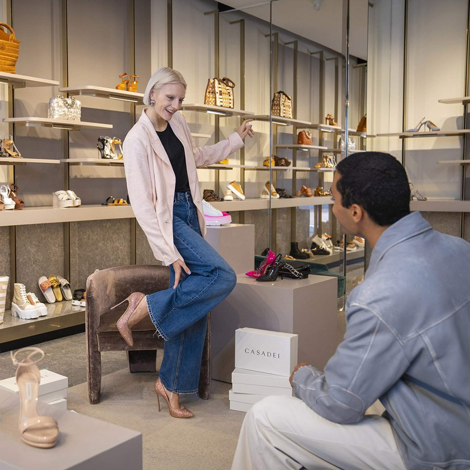Woman trying on heels in a luxury boutique at Noventa di Piave Designer Outlet, talking to a seated man.